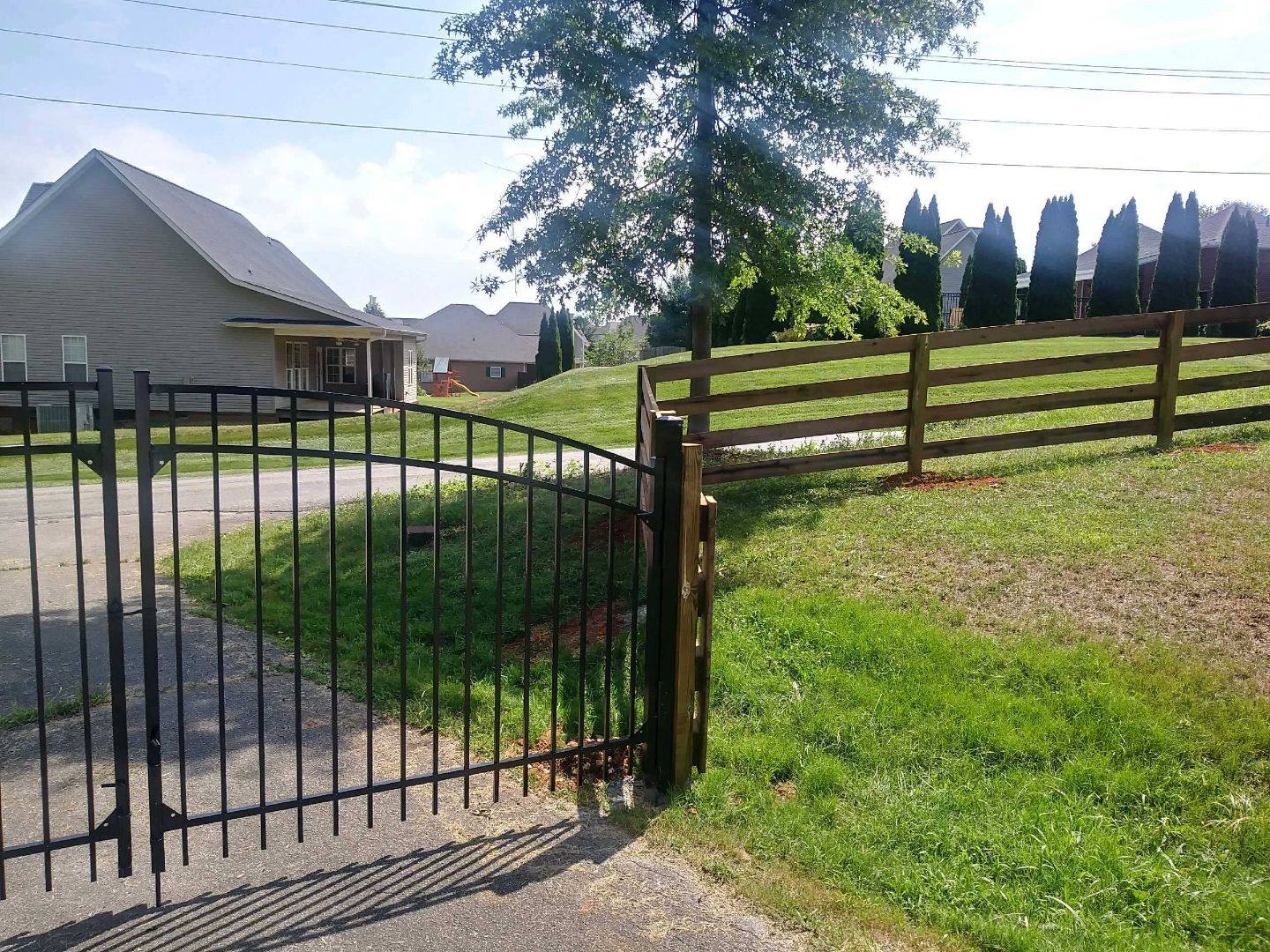 Black metal driveway gate beside a four-board wood rail fence