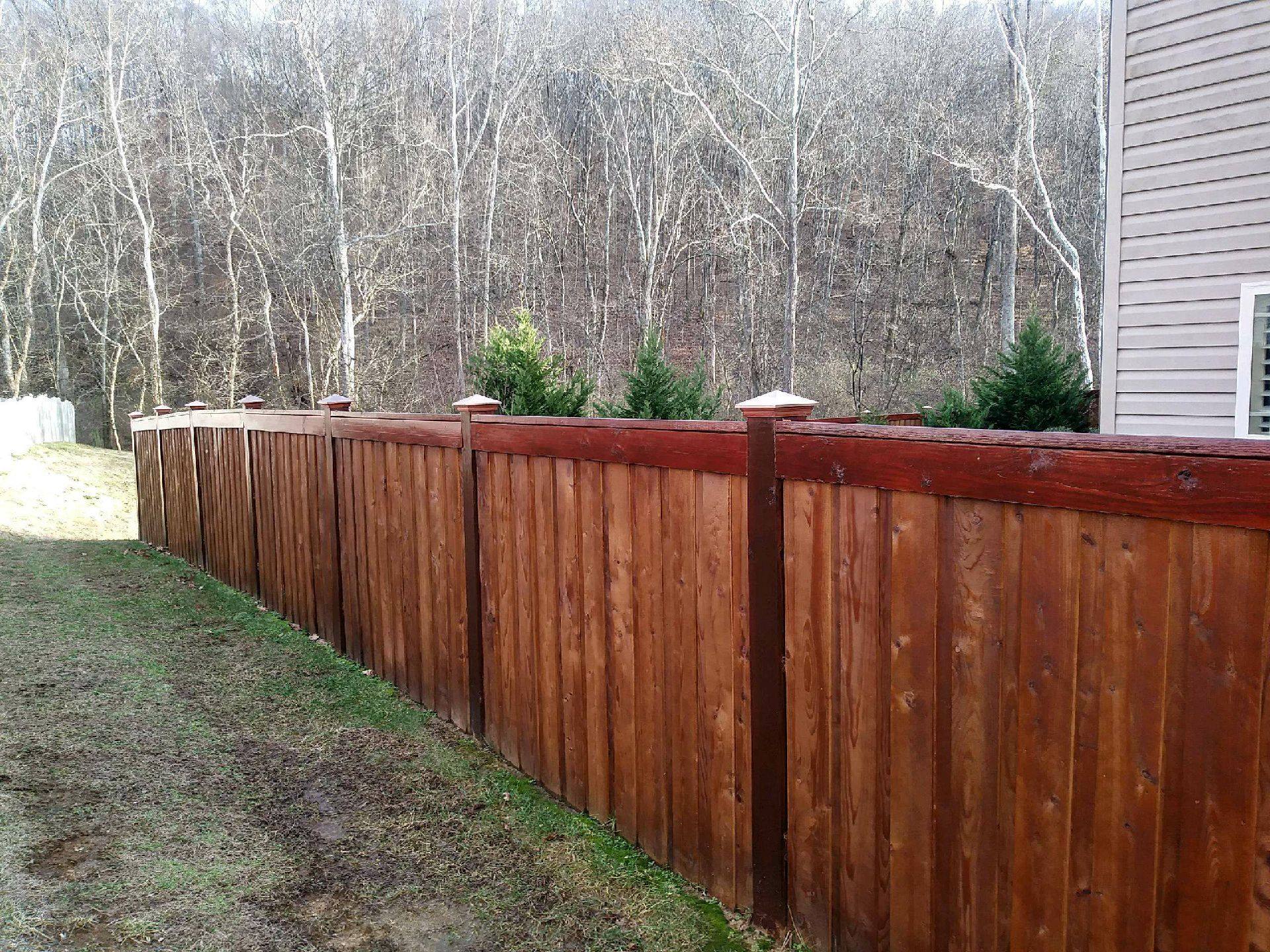 Long stained wood privacy fence with capped posts beside woods